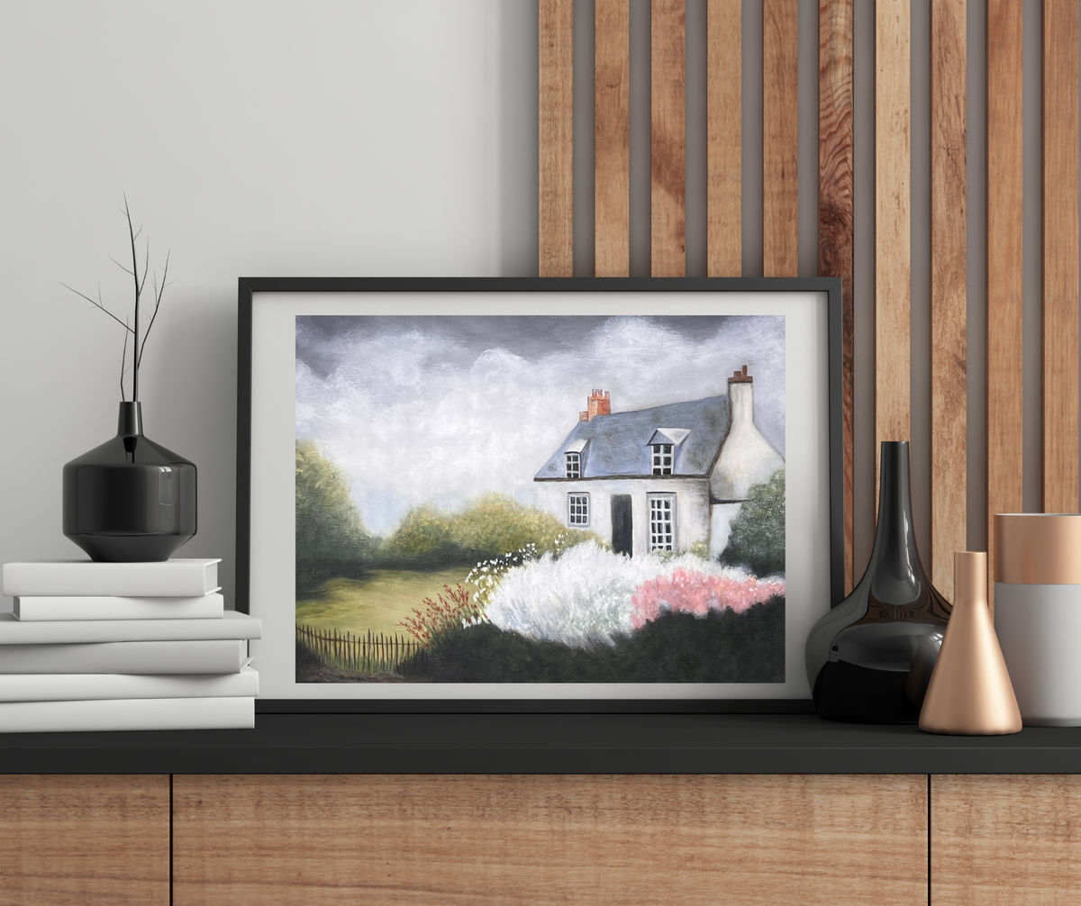 A framed landscape art print depicting a white cottage with a gray roof surrounded by green fields and pink flowers, styled on a wooden console table with decorative vases.