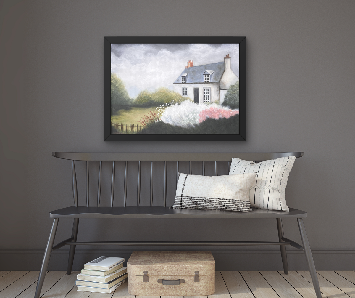 A framed landscape painting of a cottage in a grassy field with pink flowers, displayed on a dark gray wall above a gray wooden bench with pillows and a vintage suitcase.