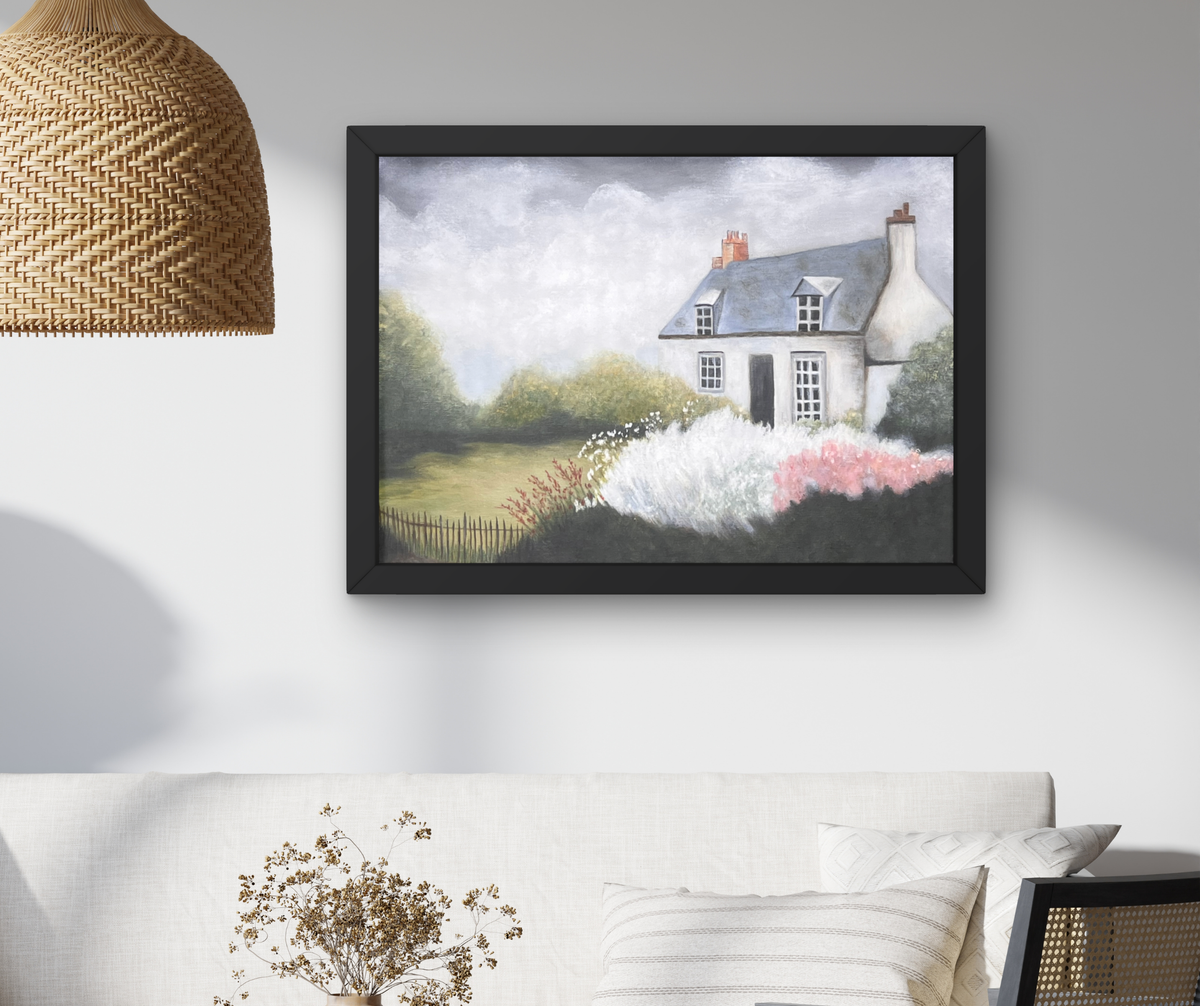 A framed landscape painting of a white cottage with pink flowers and green trees, displayed on a light gray wall above a white sofa with a woven pendant lamp partially visible on the left.