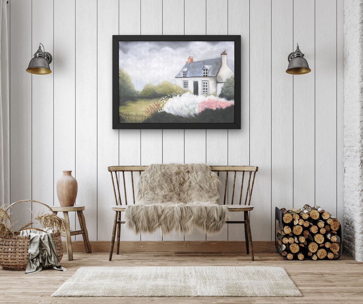 A framed landscape art print depicting a white cottage with pink flowers in the foreground, displayed on a white paneled wall above a wooden bench with a faux fur throw in a styled room setting.