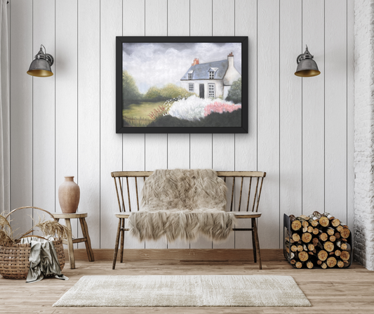 A framed landscape art print depicting a white cottage with pink flowers in the foreground, displayed on a white paneled wall above a wooden bench with a faux fur throw in a styled room setting.