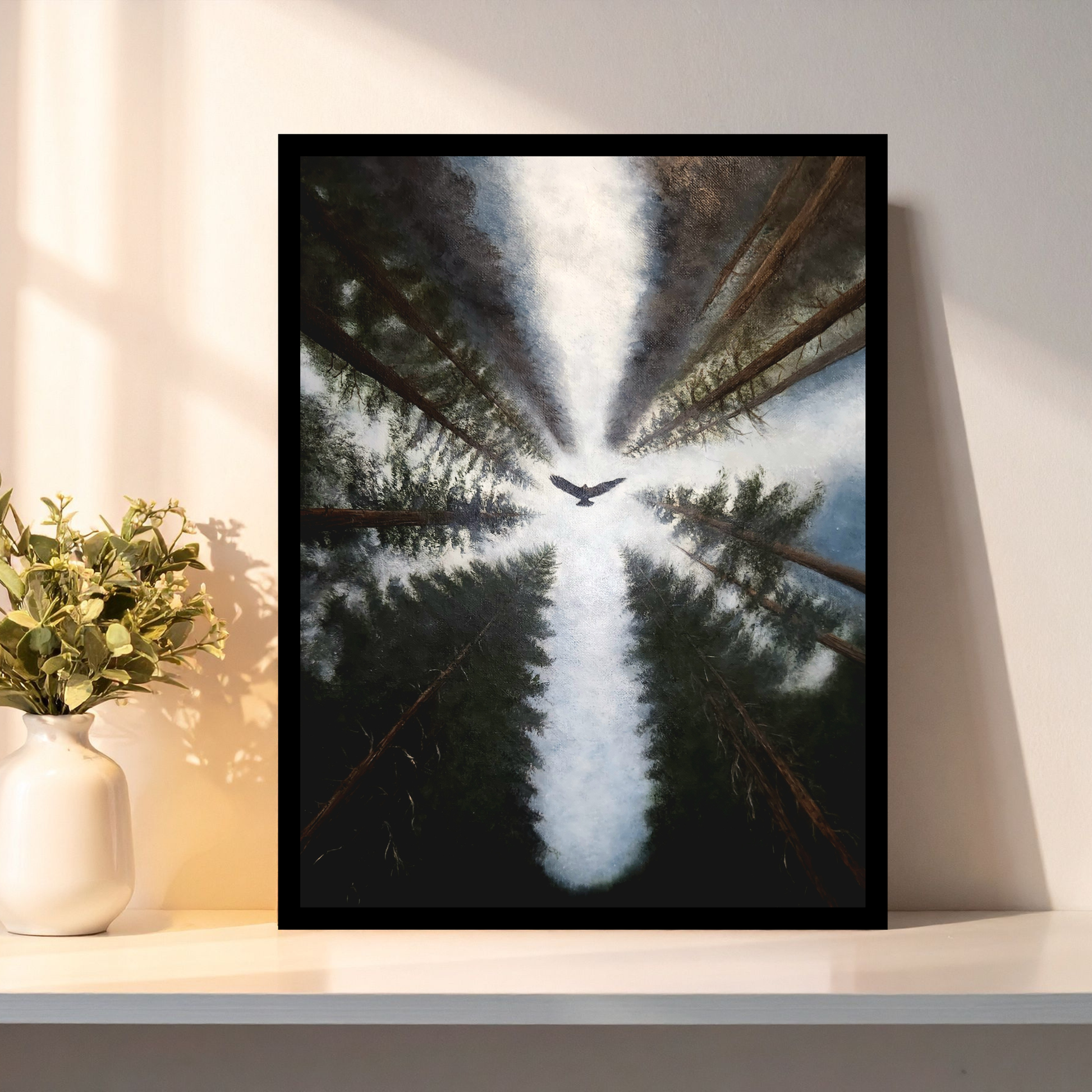 A framed vertical art print leaning against a wall on a shelf, showing an upward view of tall dark trees with a bright white light or bird at the center. A white vase with greenery sits beside it on the shelf.