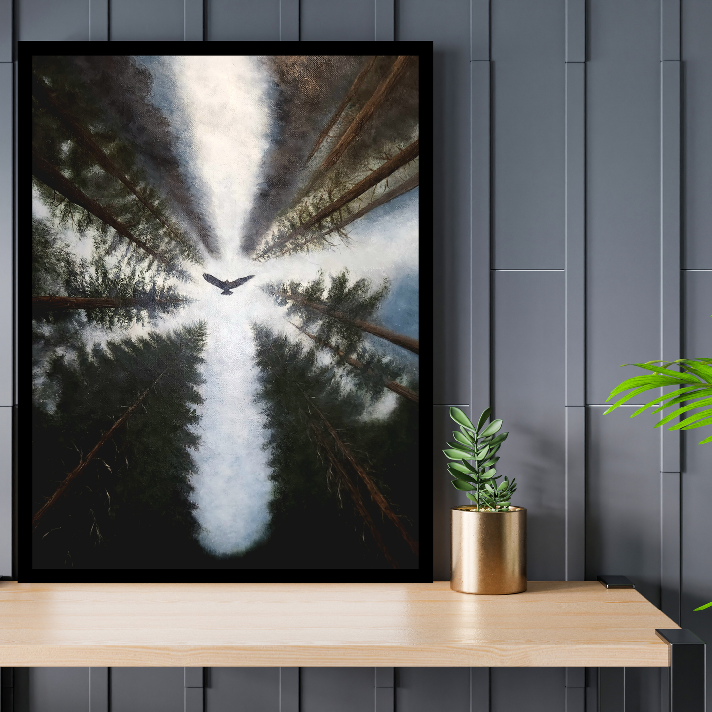 A framed art print depicting an aerial view of a forest with a bird flying toward the center under a bright light, displayed on a light wood shelf next to a potted plant against a dark gray paneled wall.