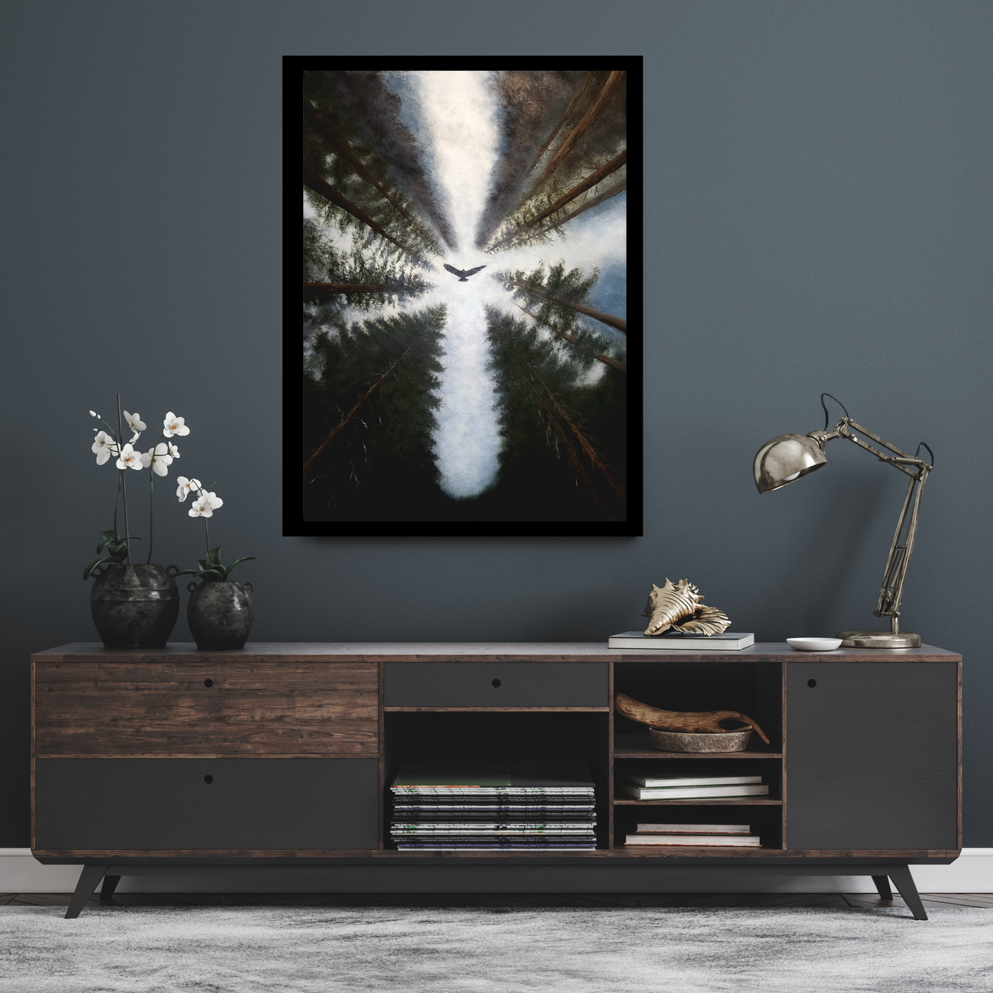 A framed vertical art print depicting a forest scene with a bright light radiating from the center, hanging on a dark gray wall above a modern wooden sideboard with decorative items.