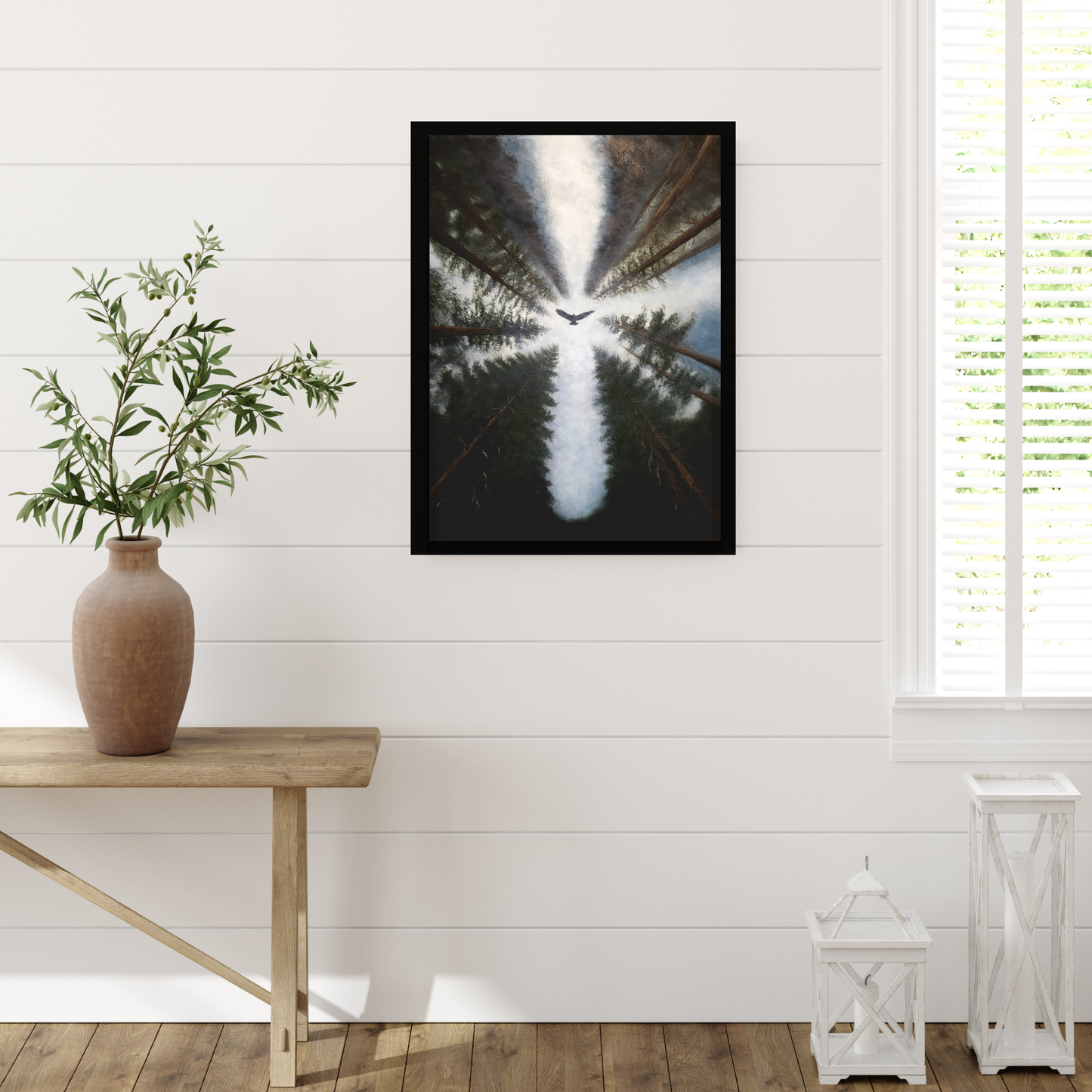 A framed vertical art print depicting a bird flying over a forest reflection in water, styled on a white shiplap wall above a wooden bench with a vase of greenery.